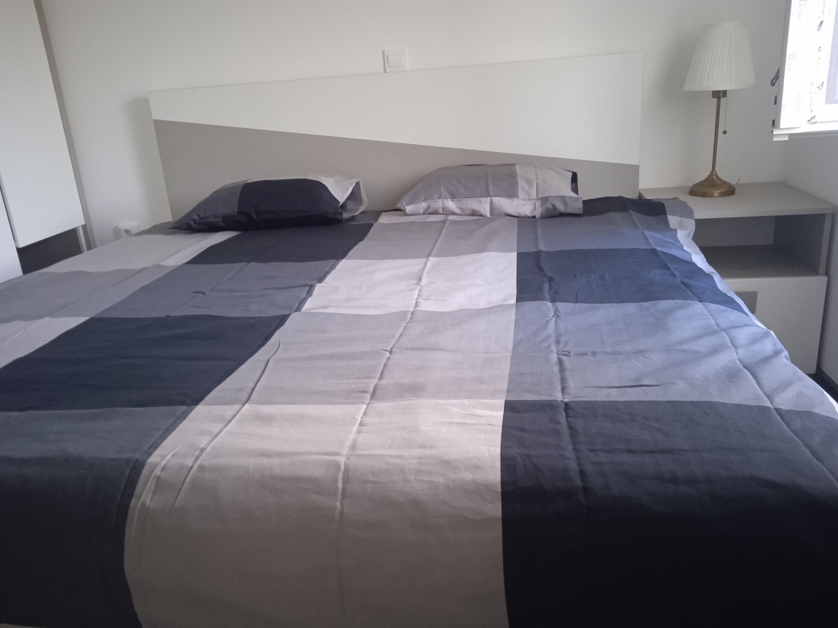 Siististi pedattu sänky, jossa on ruutukuviollinen päiväpeite, yöpöytä lampun kanssa ja ikkuna, jossa on sälekaihtimet.