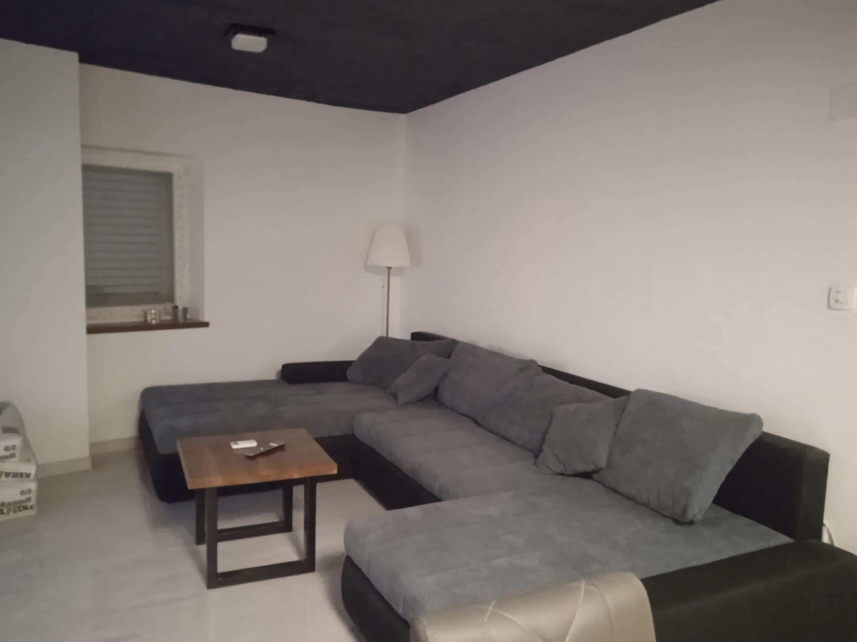 Un salon avec un canapé d’angle gris, une table basse en bois, un lampadaire et une fenêtre avec des stores.