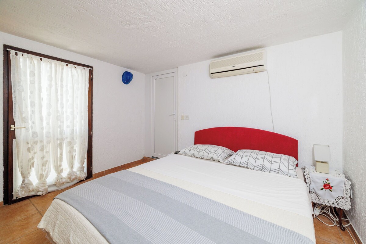Ein Schlafzimmer mit einem Bett, einer weißen Klimaanlage, einem Nachttisch mit Lampe und einer mit Vorhängen versehenen Tür.