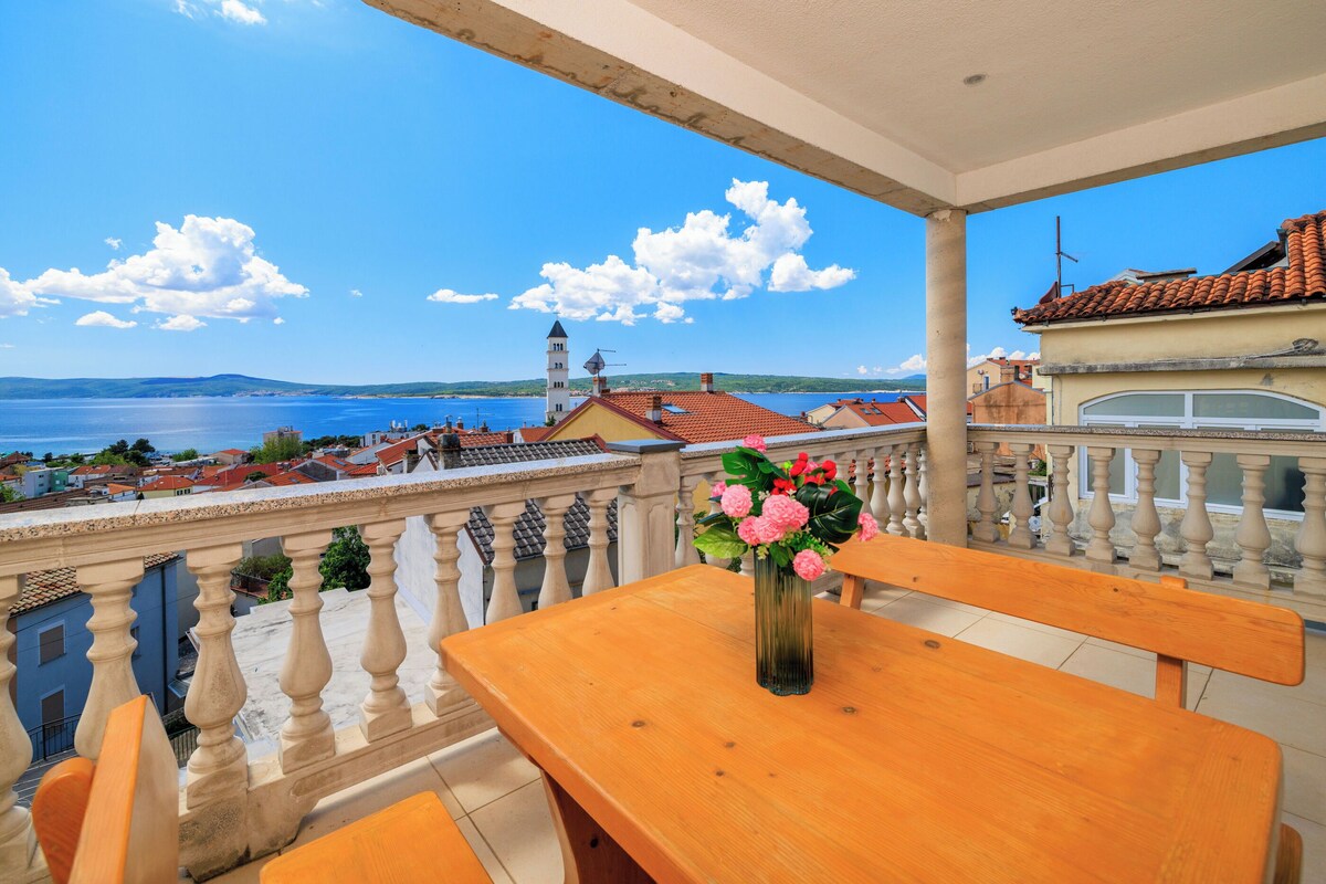 Ein Balkon mit Holztisch und -stühlen, einer Blumenvase und Blick auf das Meer und die Gebäude.