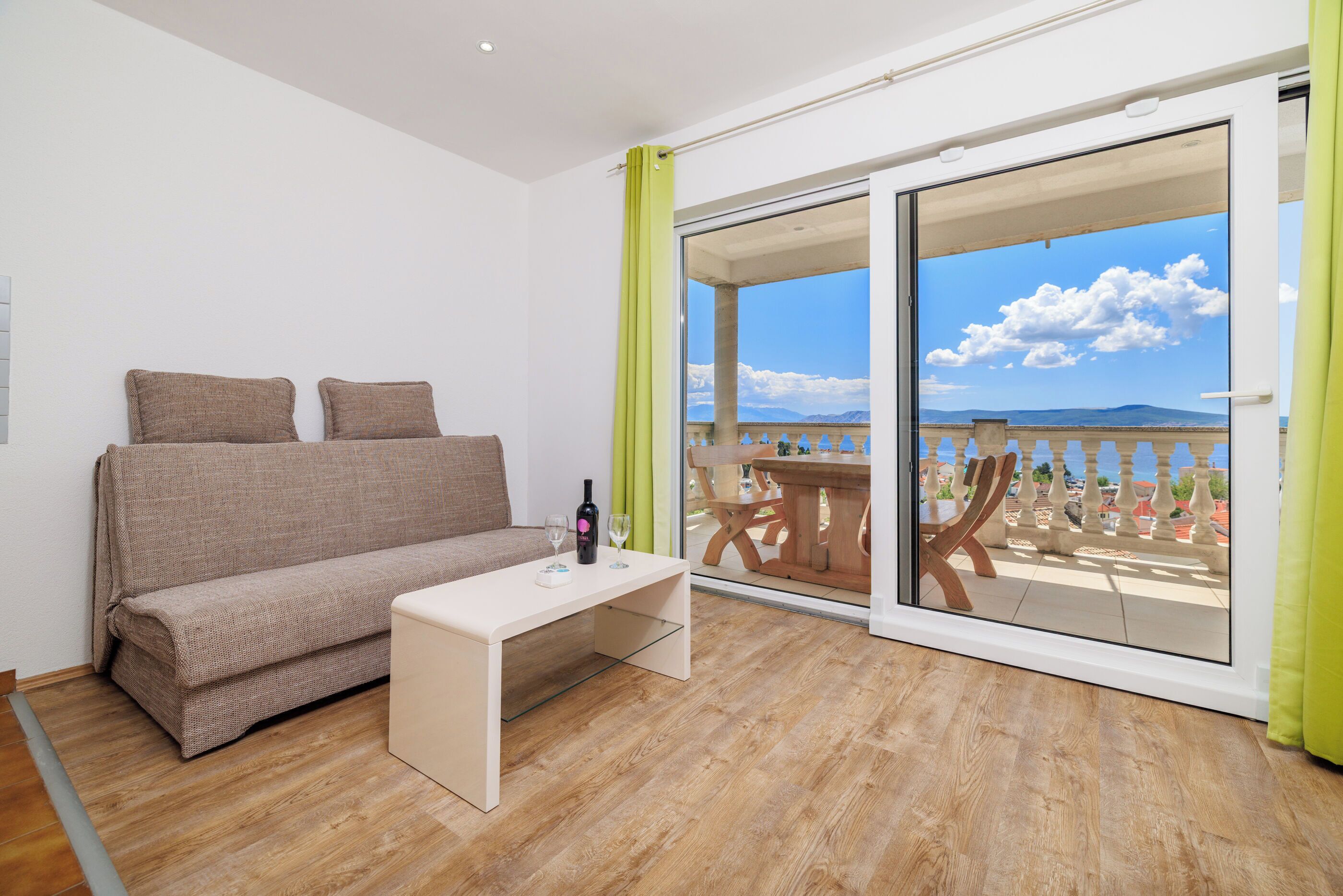 A room with a sofa, a small table, and a view of a balcony with a wooden table and chairs.