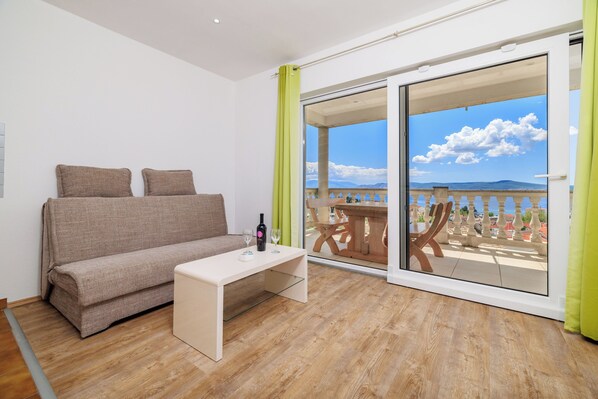 A room with a sofa, a small table, and a view of a balcony with a wooden table and chairs. - Apartments Andrijanic (Crikvenica)