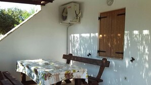 Un espace extérieur abrité, aménagé pour prendre les repas, avec une table pour deux personnes, un banc en bois et un climatiseur fixé au mur.