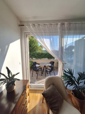 A room with a wooden dresser, a chair, a window with sheer curtains, and a view of an outdoor seating area with chairs and greenery. - Apartment Goran (Makarska)