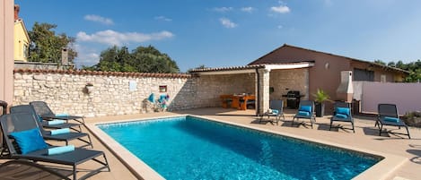A swimming pool with lounge chairs, a stone wall, and a covered patio area.