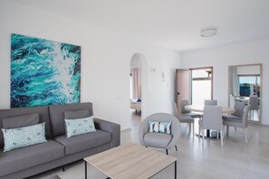 A modern living room with a grey sofa, a coffee table, and a large abstract painting. There is a dining area with a table and chairs. - Villa Montane 10 - Three Bedroom Villa, Sleeps 6 (Playa Blanca)