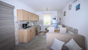 A compact kitchen with wooden cabinets, a microwave, and a sink. There is a small dining area with a table and chairs. A bed with pillows is visible in the foreground. - Apartments Plavsic (Novi Vinodolski)