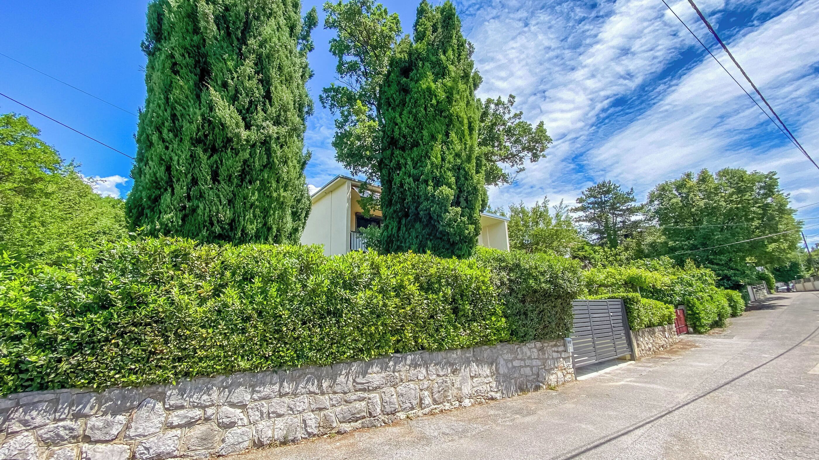 A residential area with stone walls, tall trees, and a paved road.