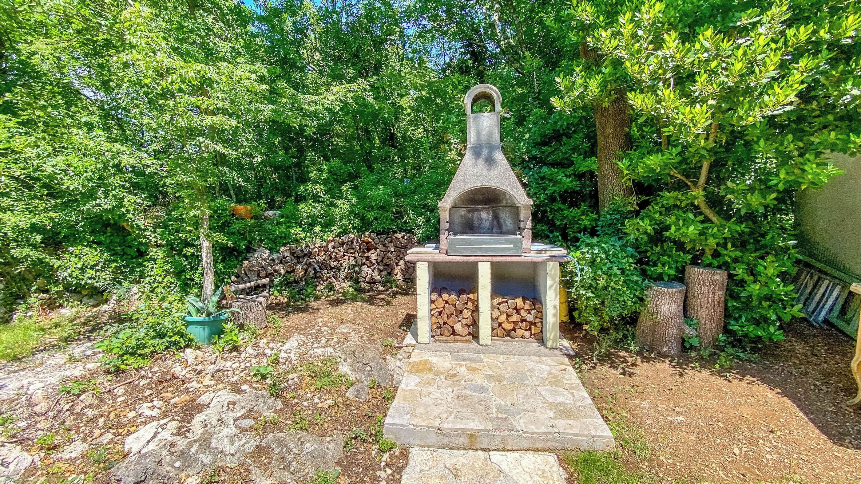 A stone-built outdoor oven with a chimney, surrounded by a garden with trees and a stone pathway.
