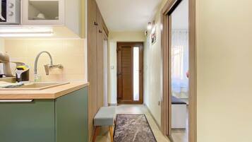 A narrow hallway with a kitchen area on the left, a wooden door at the end, and a bench in the center.