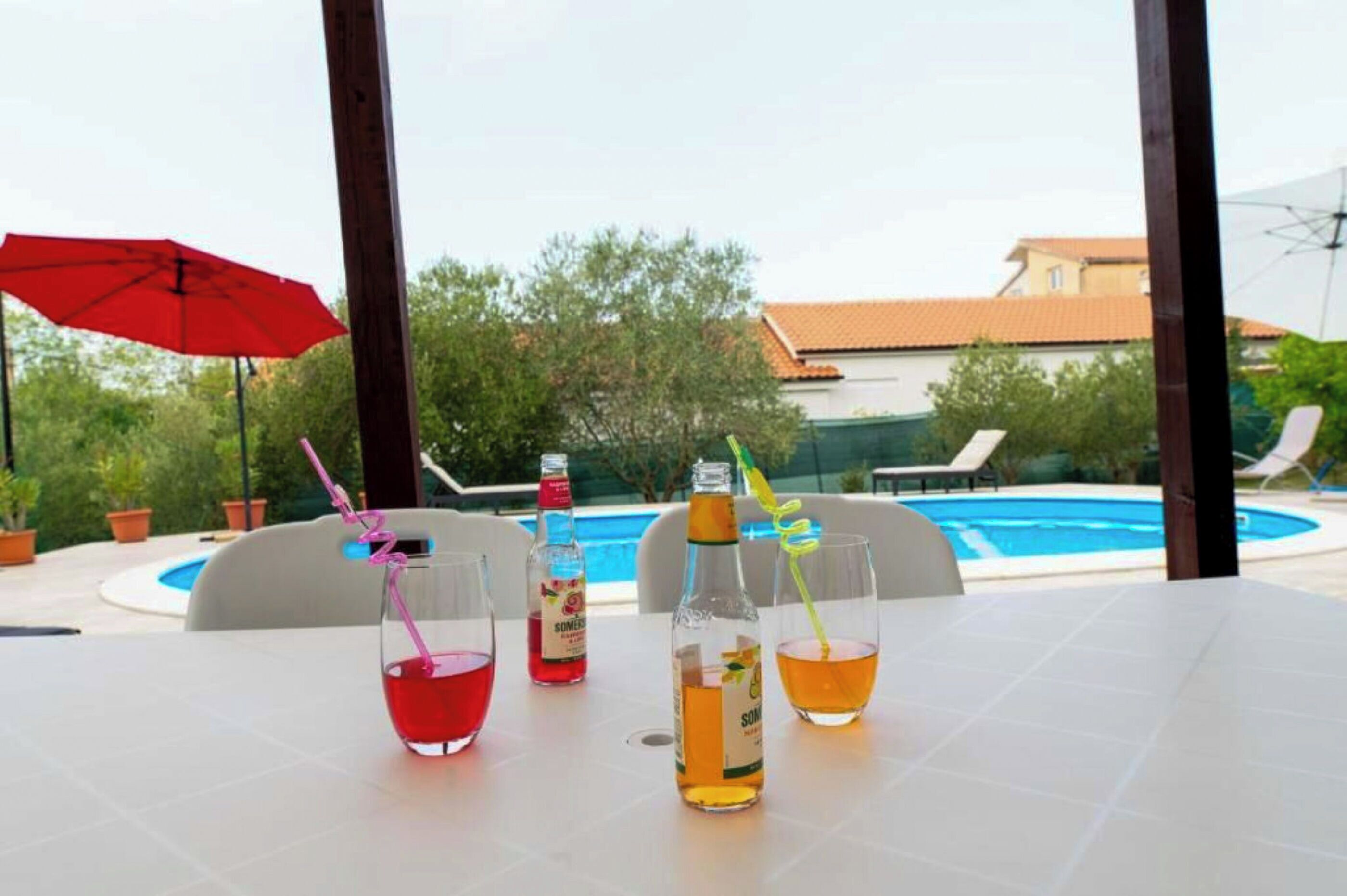 Une table avec des boissons et un parasol rouge, une piscine et des chaises longues à l’arrière-plan.