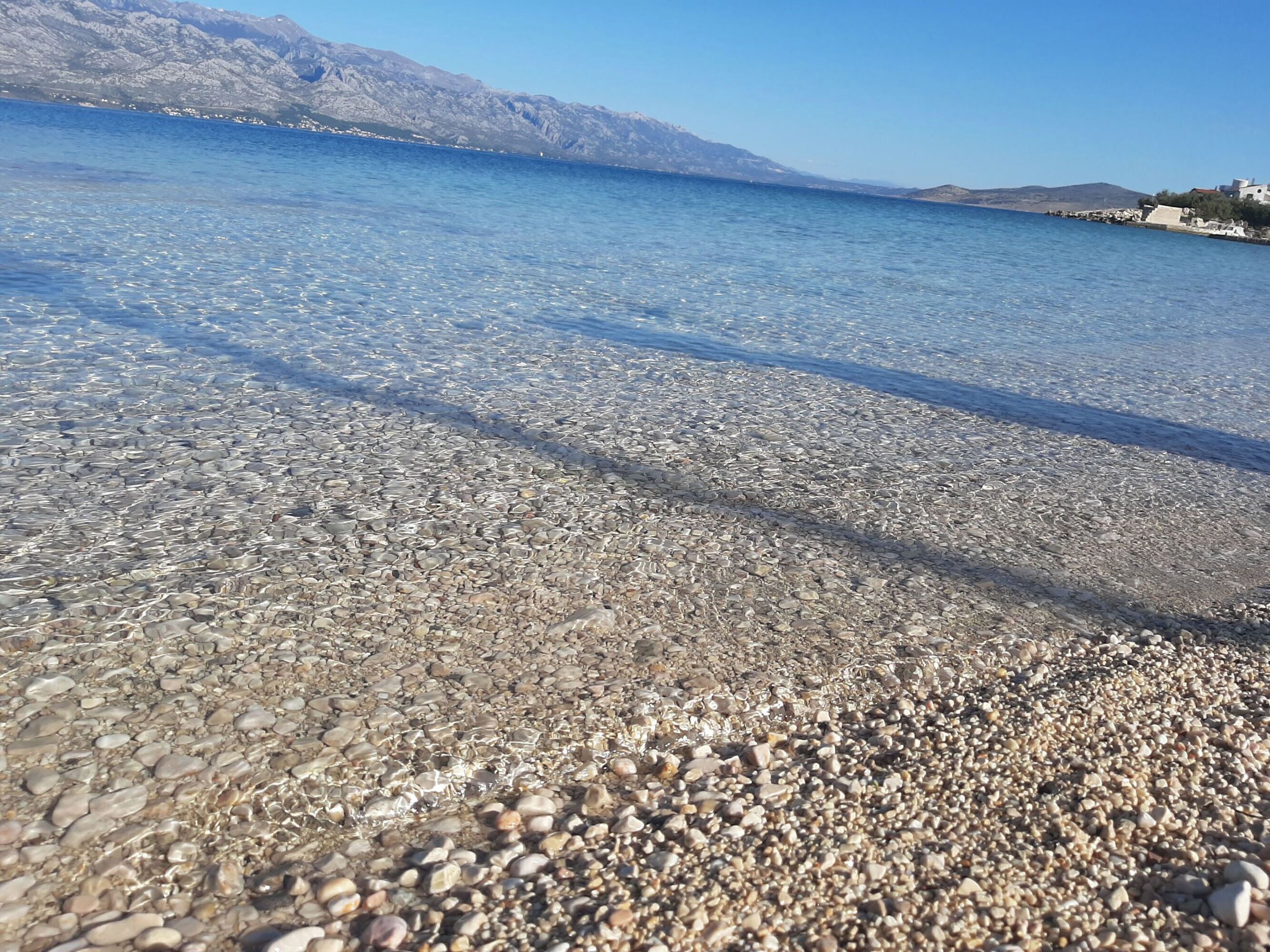 Una playa de guijarros con aguas cristalinas y montañas al fondo.