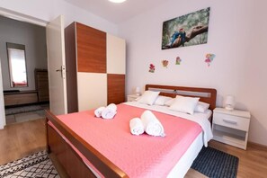 Ein ordentlich bezogenes Bett mit einer rosafarbenen Tagesdecke und weißen Kissen, ein hölzerner Kleiderschrank, ein Nachttisch mit Lampe und ein Bild an der Wand.