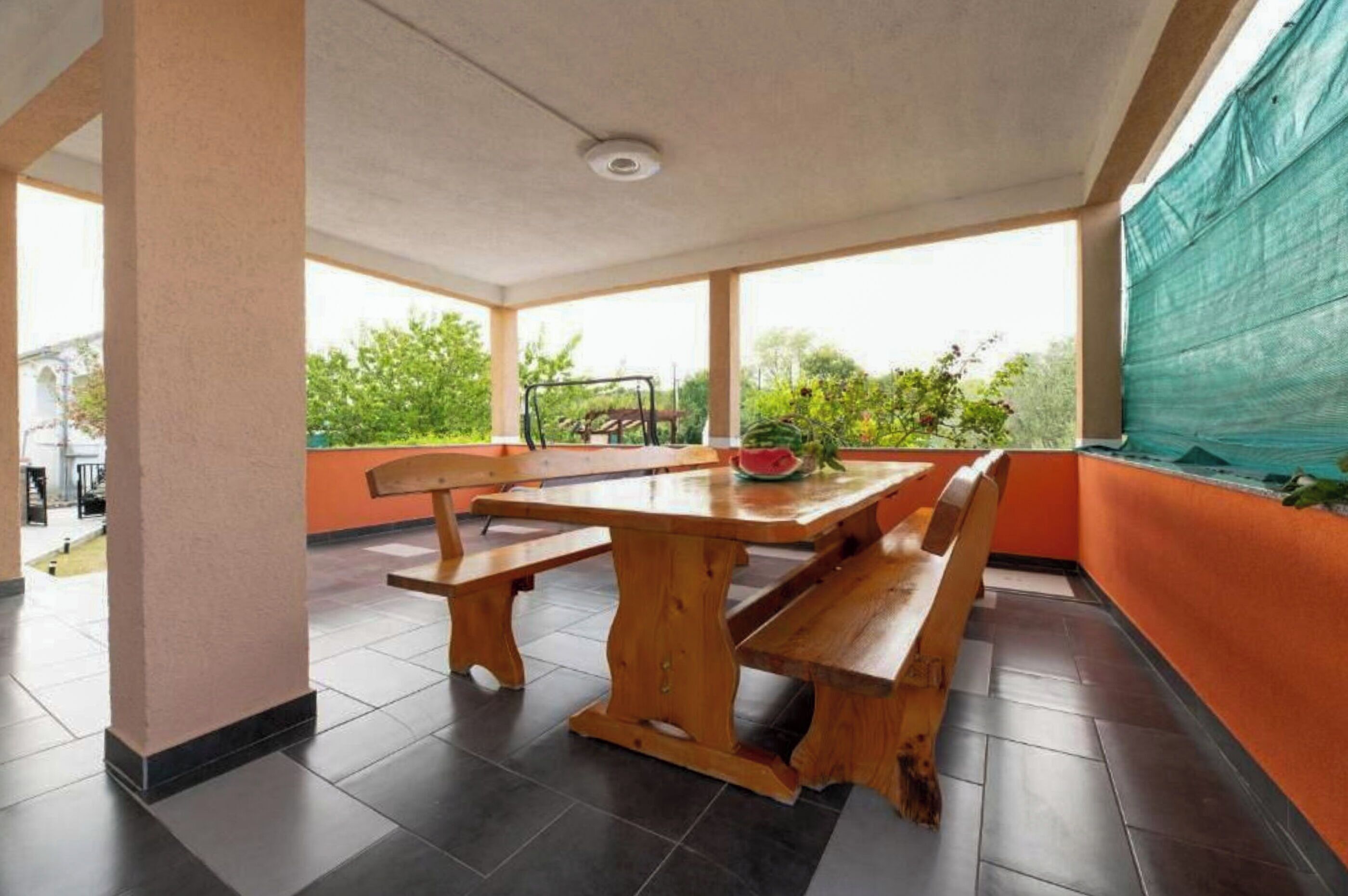 A covered patio area with a wooden table and benches, a view of greenery outside, and a ceiling light fixture.