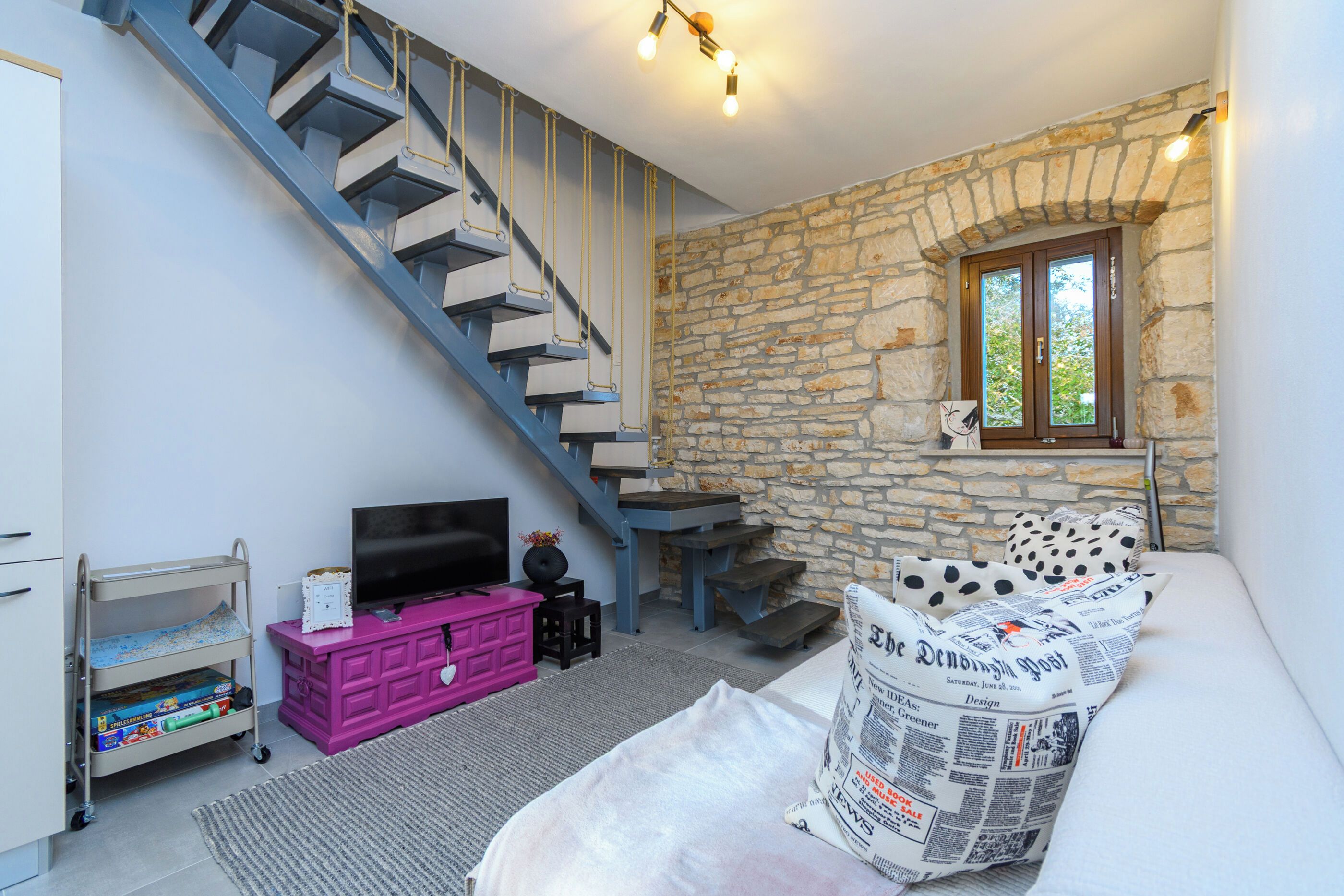 A room with a staircase, a television, a pink cabinet, and a window with a newspaper.