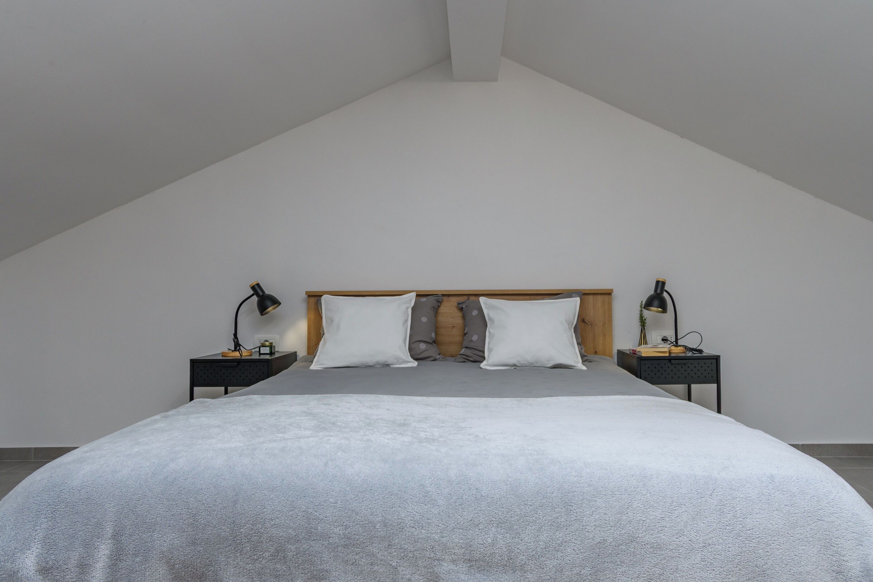 Un dormitorio con techo inclinado, una cama grande con edredón gris, dos mesitas de noche con lámparas y un cabecero de madera.