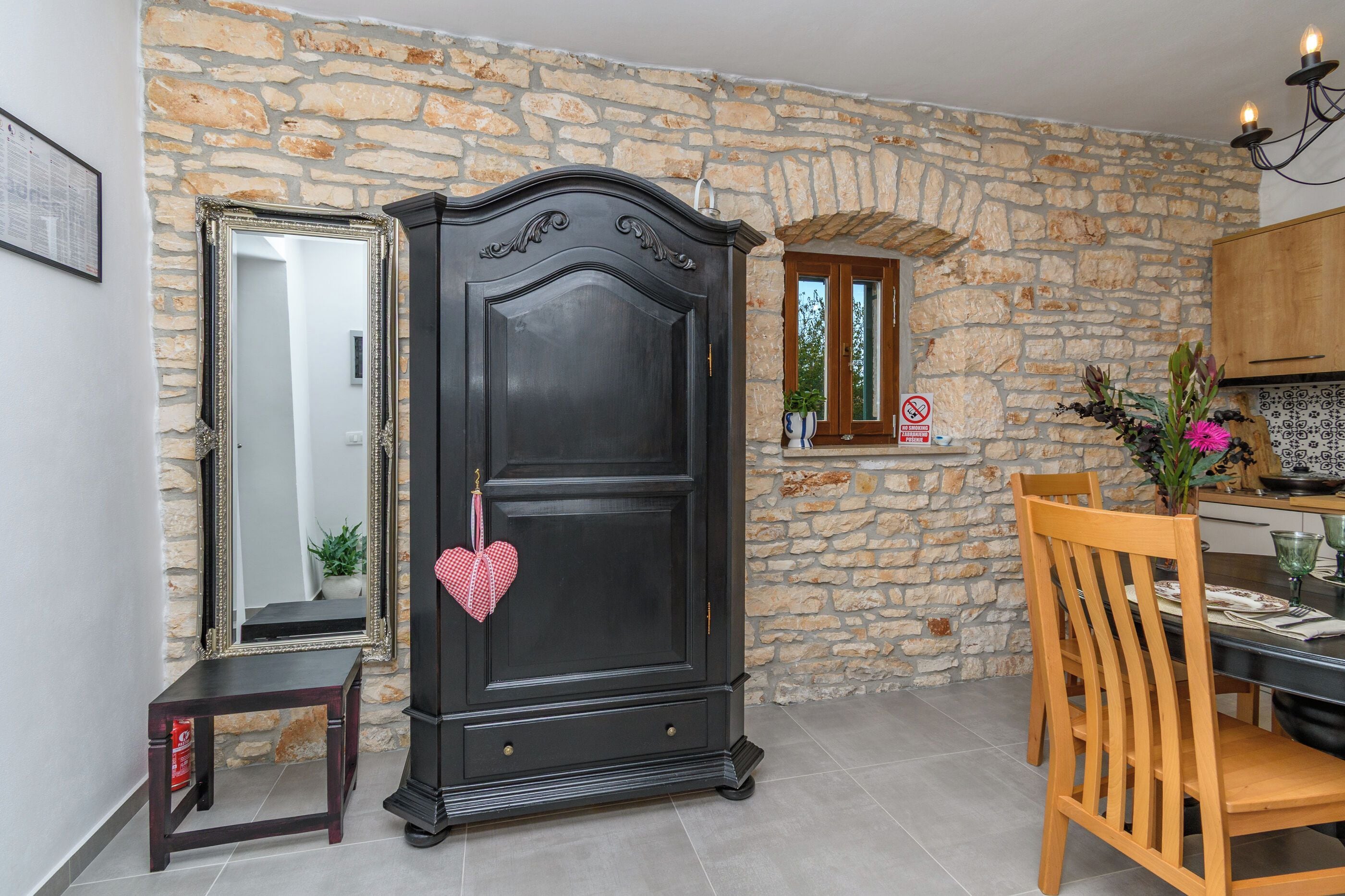 A room with a stone wall, a dark wooden wardrobe, a mirror, a dining table with chairs, and a kitchen area.