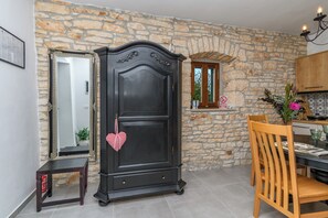 A room with a stone wall, a dark wooden wardrobe, a mirror, a dining table with chairs, and a kitchen area.