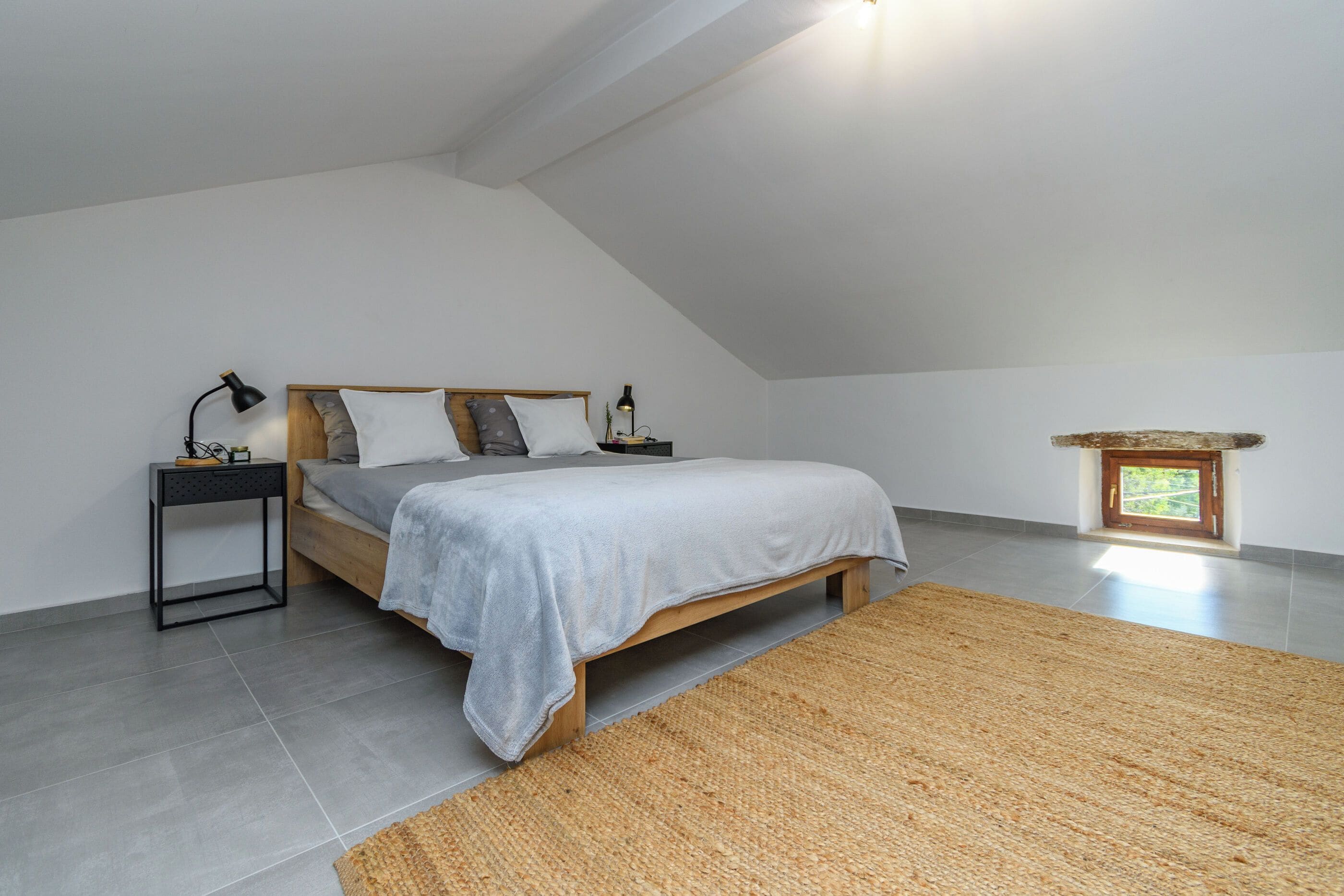 Une chambre avec un plafond en pente, un lit recouvert d’une couverture grise, deux tables de chevet avec des lampes et une petite fenêtre.