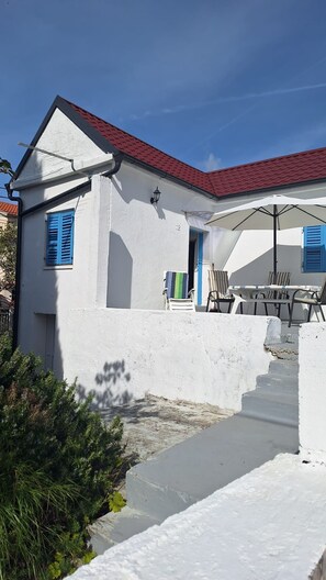 A white building with a red roof, blue shutters, and a staircase leading to a covered outdoor area with chairs and a table. - House House Marijan (Karlobag)