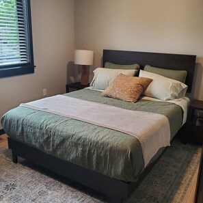 A bed with a green bedspread and pillows, a bedside lamp, and a window with blinds. - Newly Constructed 2 bedroom, 1 bath home, quiet, private. (Shelton)