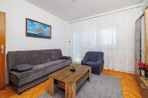 A living room with a grey sofa, a wooden coffee table, a blue armchair, a painting of a sailboat, and a large window with sheer curtains. - Apartments Smiljka (Crikvenica)