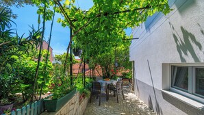 Un patio empedrado con mesa y sillas, rodeado de follaje y una pared blanca.