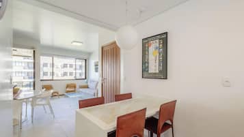 Un comedor moderno con una mesa blanca y sillas marrones, un ventanal con persianas y un cuadro en la pared.