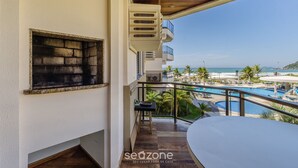 A balcony with a view of a pool and beach, a small table, and a window with a grill.
