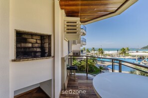 A balcony with a view of a pool and beach, a small table, and a window with a grill. - Charm and Elegance by the Sea! Rit0201 (Florianópolis)