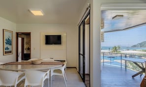 Un espace de repas moderne avec une table en verre et des chaises blanches, un téléviseur à écran plat fixé au mur, et une vue sur une piscine et des montagnes, grâce à de grandes fenêtres.