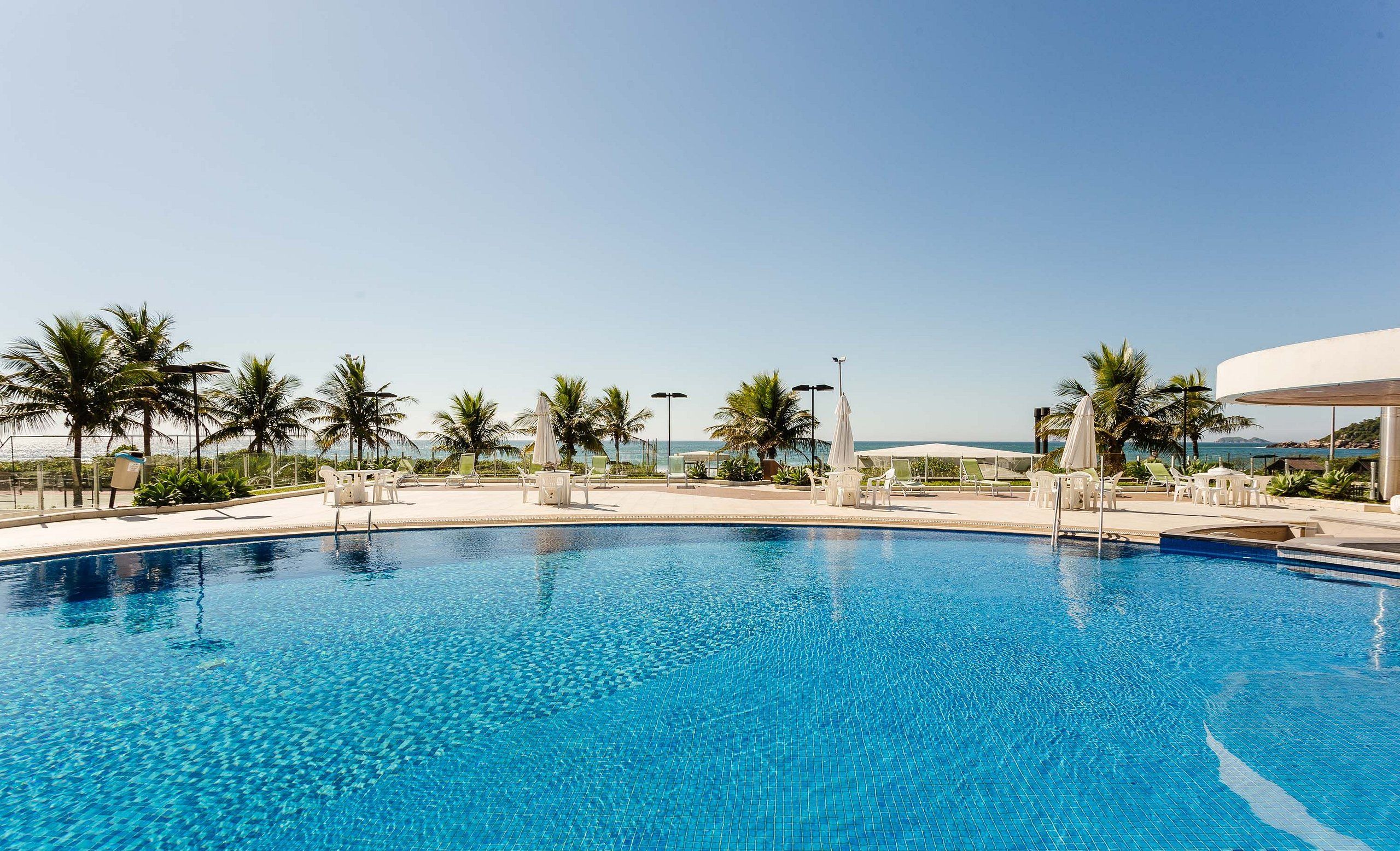Une piscine d’un bleu éclatant, entourée de chaises longues blanches et de parasols, avec des palmiers et une vue sur la mer.
