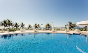 En klar, blå pool omgivet af hvide liggestole og parasoller, med palmer og udsigt til havet.