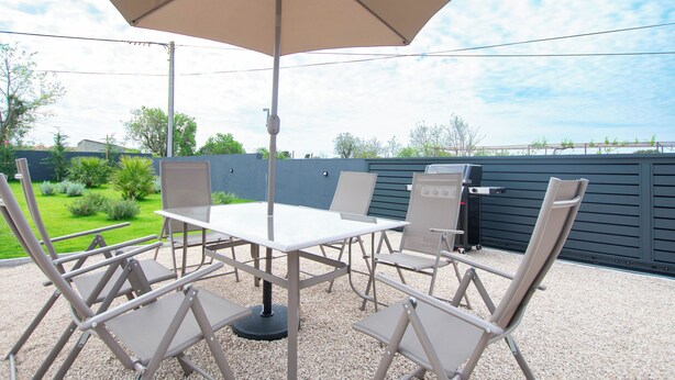 An outdoor patio with a table and chairs, a large umbrella, and a view of a garden and fence. - House Nadalina (Vrsi)