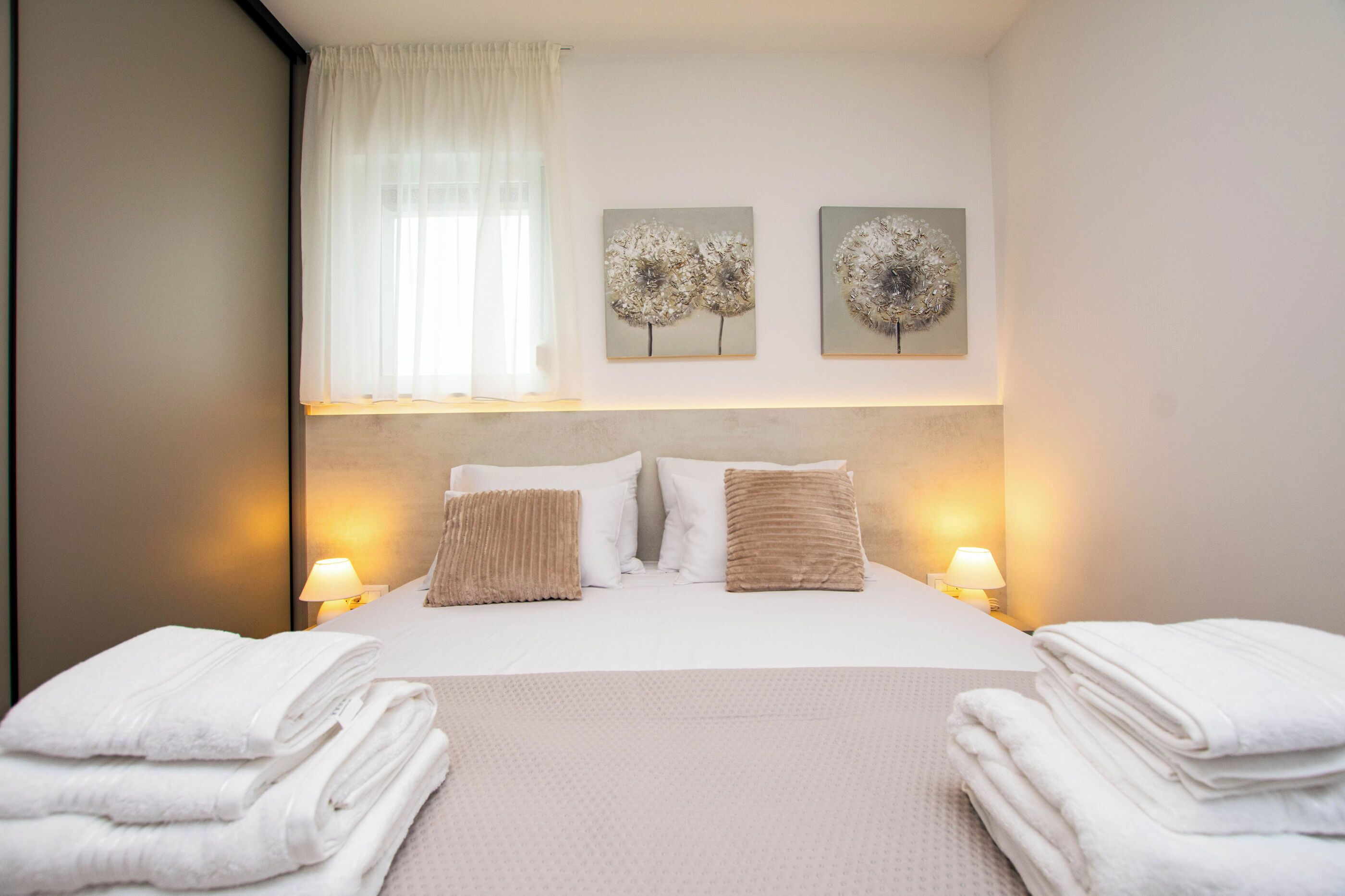 A hotel room with a bed, two wall-mounted lamps, a window with sheer curtains, and two wall art pieces.