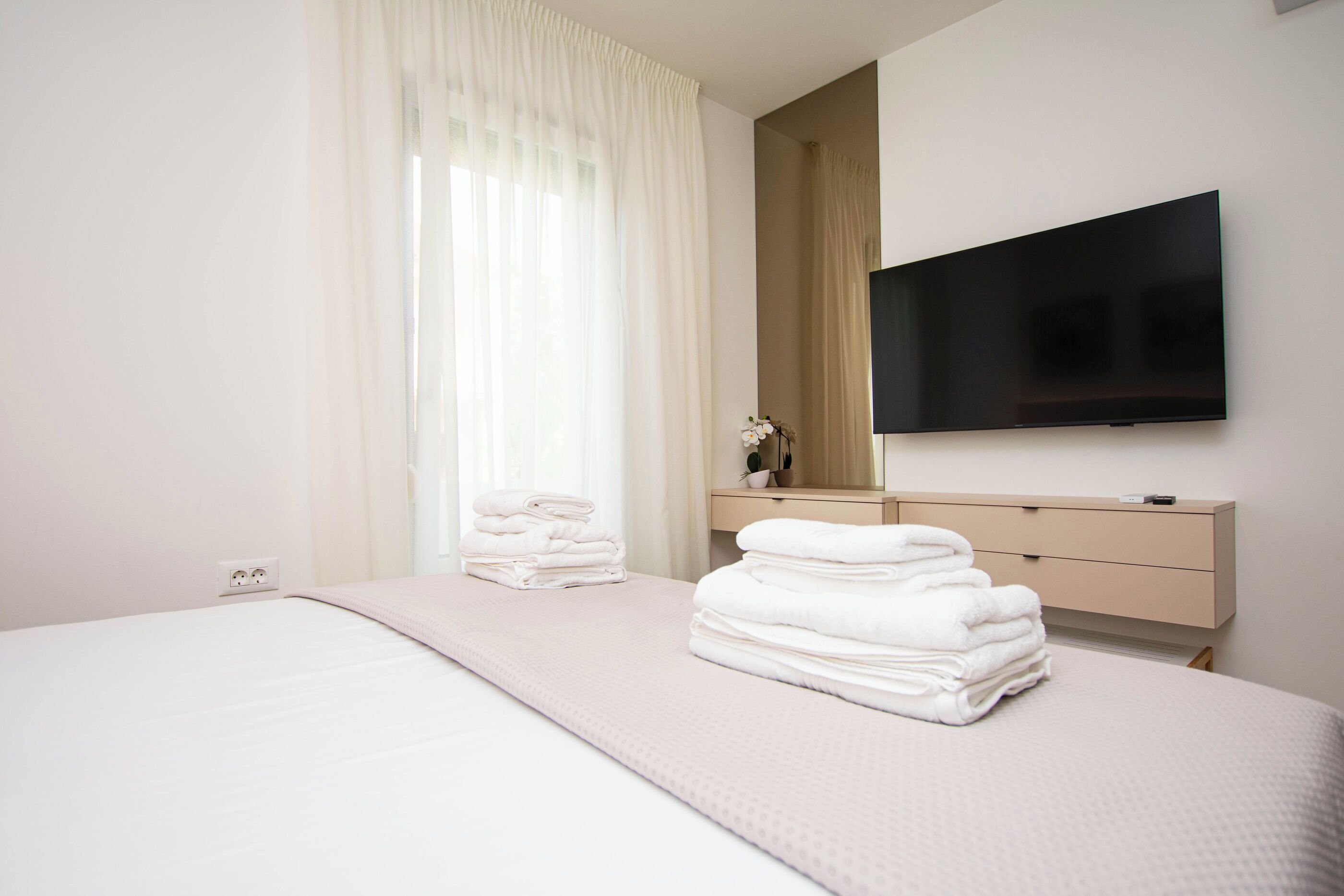 Ein Hotelzimmer mit Bett, Wandfernseher, Nachttisch mit Spiegel und Fenster mit Vorhängen.