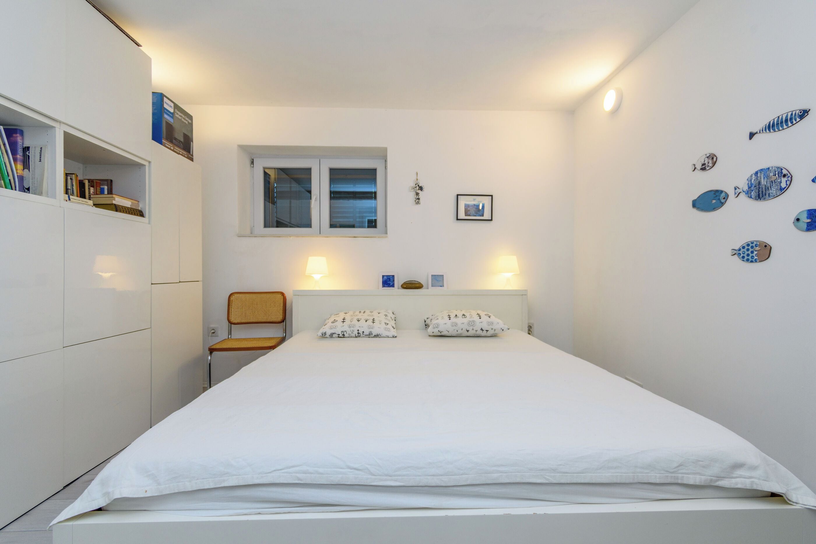 Ein schlicht eingerichtetes Schlafzimmer mit einem Bett, einem Sessel, einem Bücherregal, einem Fenster und Wanddekorationen.