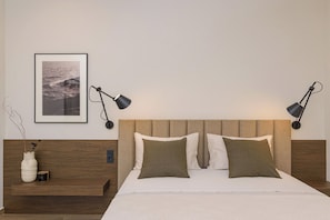 Una cama bien hecha con cabecera, dos lámparas de noche, un cuadro en la pared y una mesita de noche con un florero y una vela.