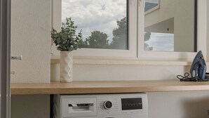 A compact laundry area with a front-loading washing machine, a window with a view of the sky, and a small shelf with a potted plant and an iron. - Apartment Azure Pine (Okrug)