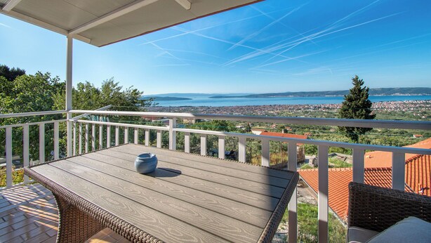 A covered outdoor area with a table and chairs, overlooking a scenic view of a coastal town and the sea. - Apartment Panorama Nicolas (Kastela)