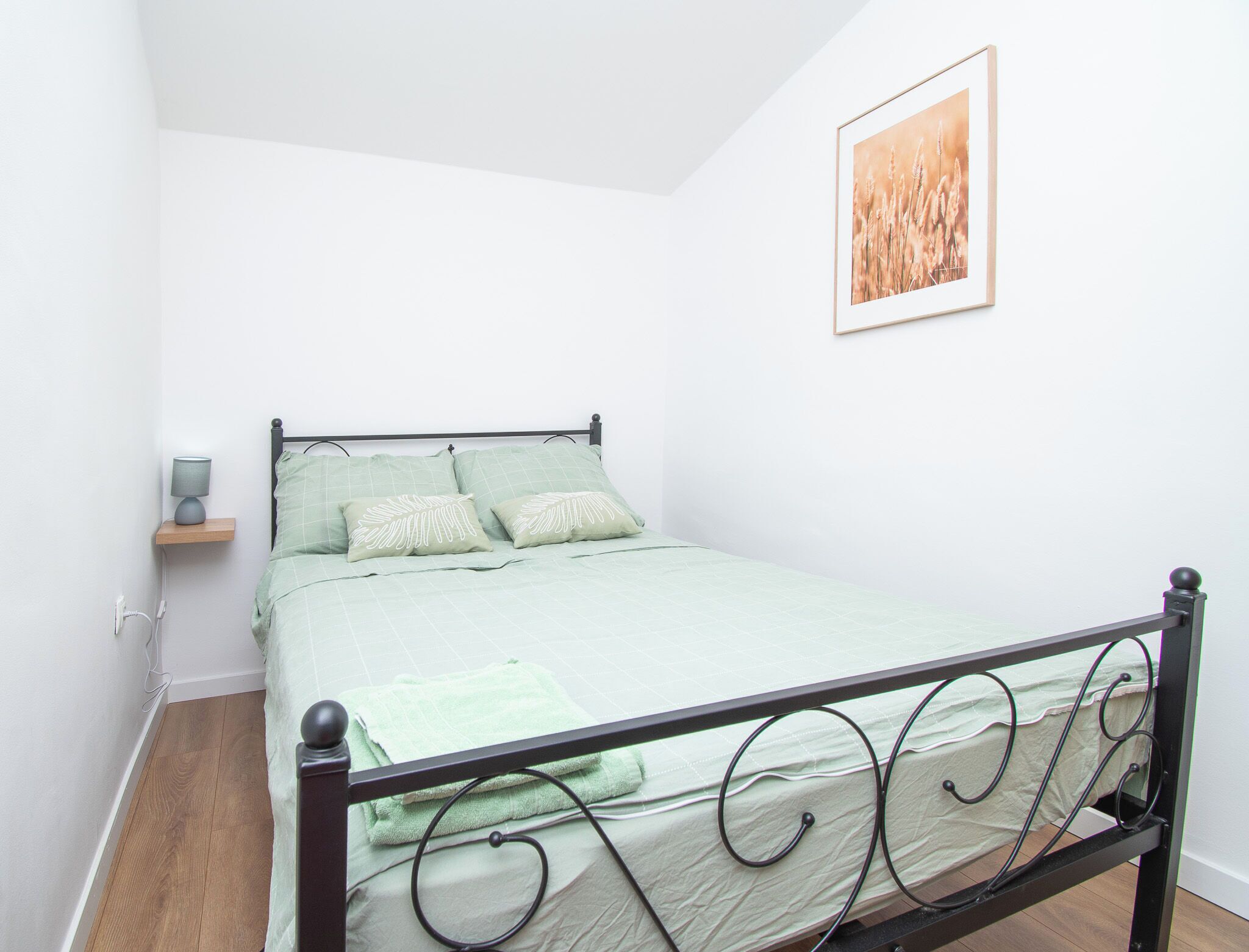 A single bed with a metal frame, a green bedspread, two pillows with leaf patterns, a bedside table with a lamp, and a framed picture on the wall.