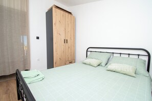 A bedroom with a bed, a wooden wardrobe, and a window with a sheer curtain. - Apartment Panorama Nicolas (Kastela)