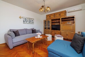 Un soggiorno con un divano, una poltrona blu, un tavolino in legno, una TV e dei mobili contenitori in legno.