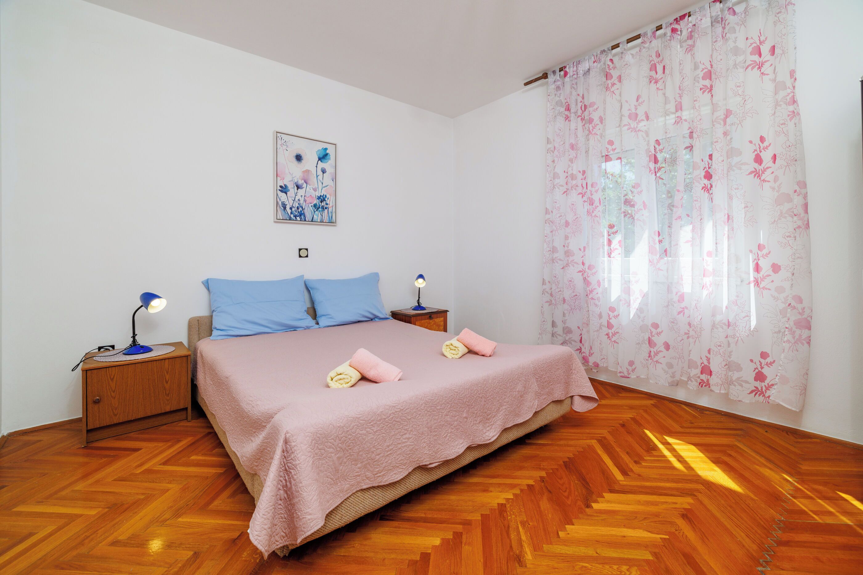 Une chambre à coucher comprenant un lit, deux tables de chevet, une lampe de chevet, un tableau accroché au mur et une fenêtre ornée de rideaux à motifs floraux.