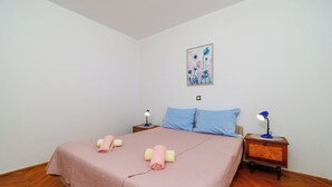 Een slaapkamer met een bed, twee kussens, een nachtkastje met een lamp en een aan de muur hangend schilderij.