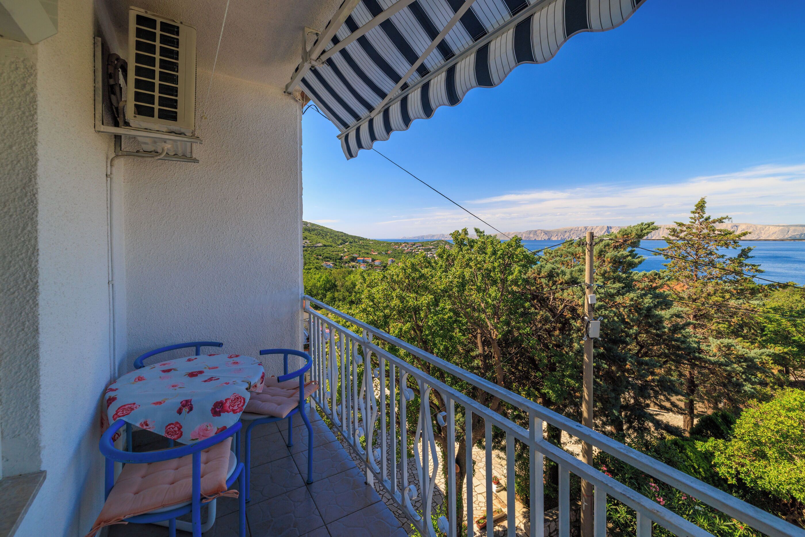 Ein Balkon mit Tisch und Stühlen, einem gestreiften Sonnensegel und Blick auf eine Küstenlandschaft.