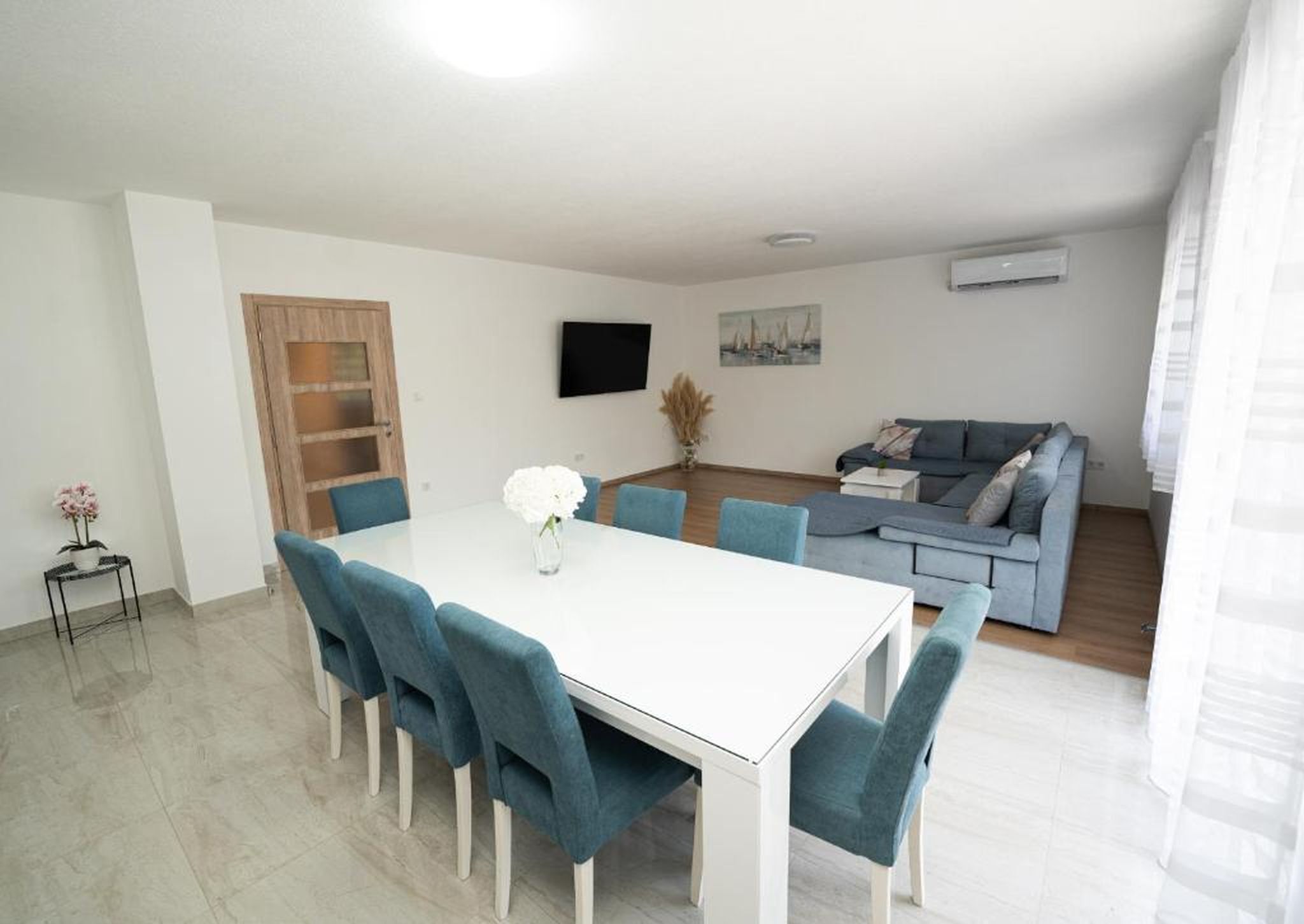 Una sala de estar moderna con una mesa de comedor blanca, sillas azules, un sofá gris y un televisor de pantalla plana en la pared.