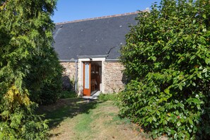 A stone building with a dark tiled roof, a wooden door, and a small garden area with grass and shrubs.