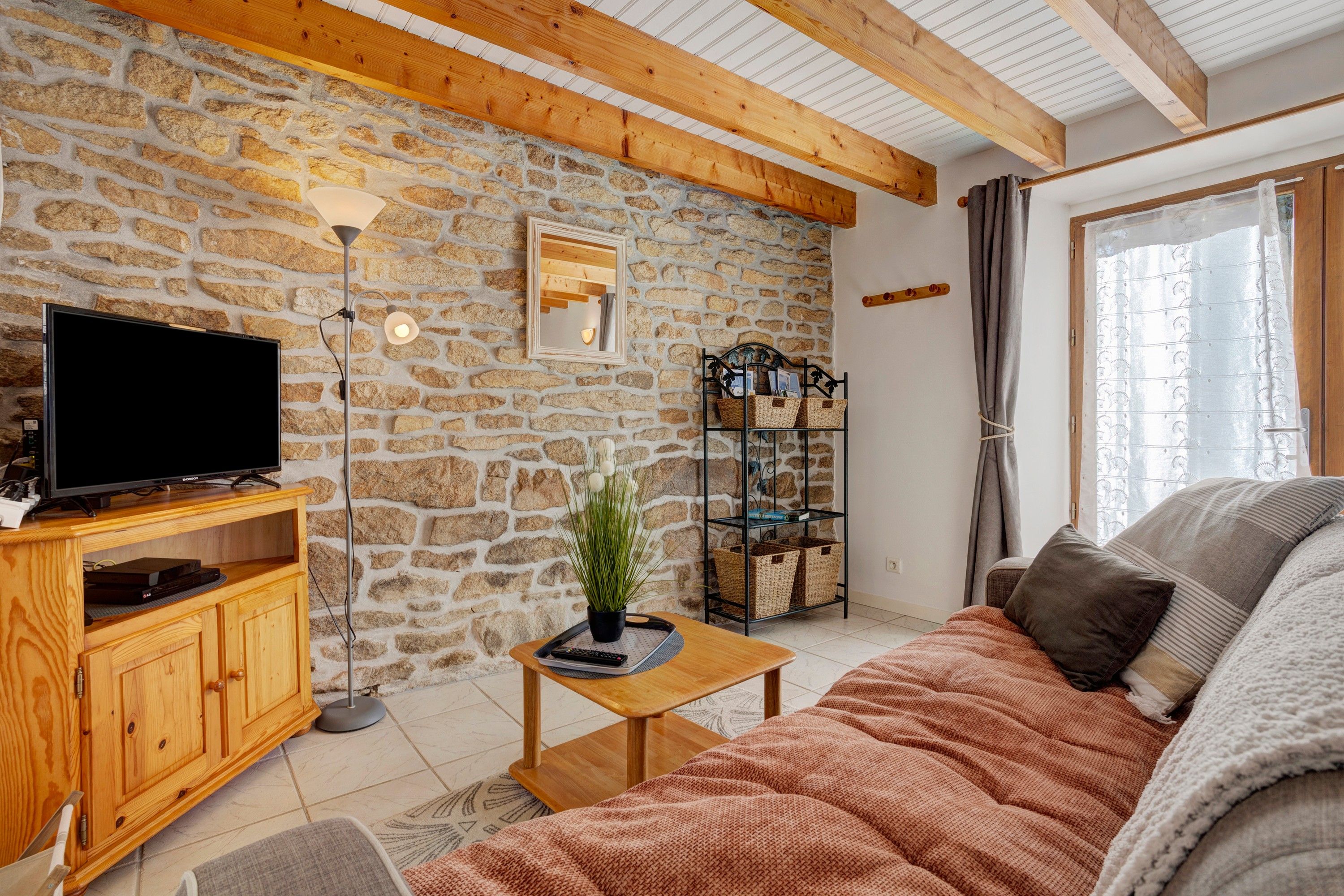 Un salón acogedor con pared de piedra, vigas de madera, un televisor de pantalla plana, un aparador de madera, una mesa de centro, una estantería y una ventana con cortinas.