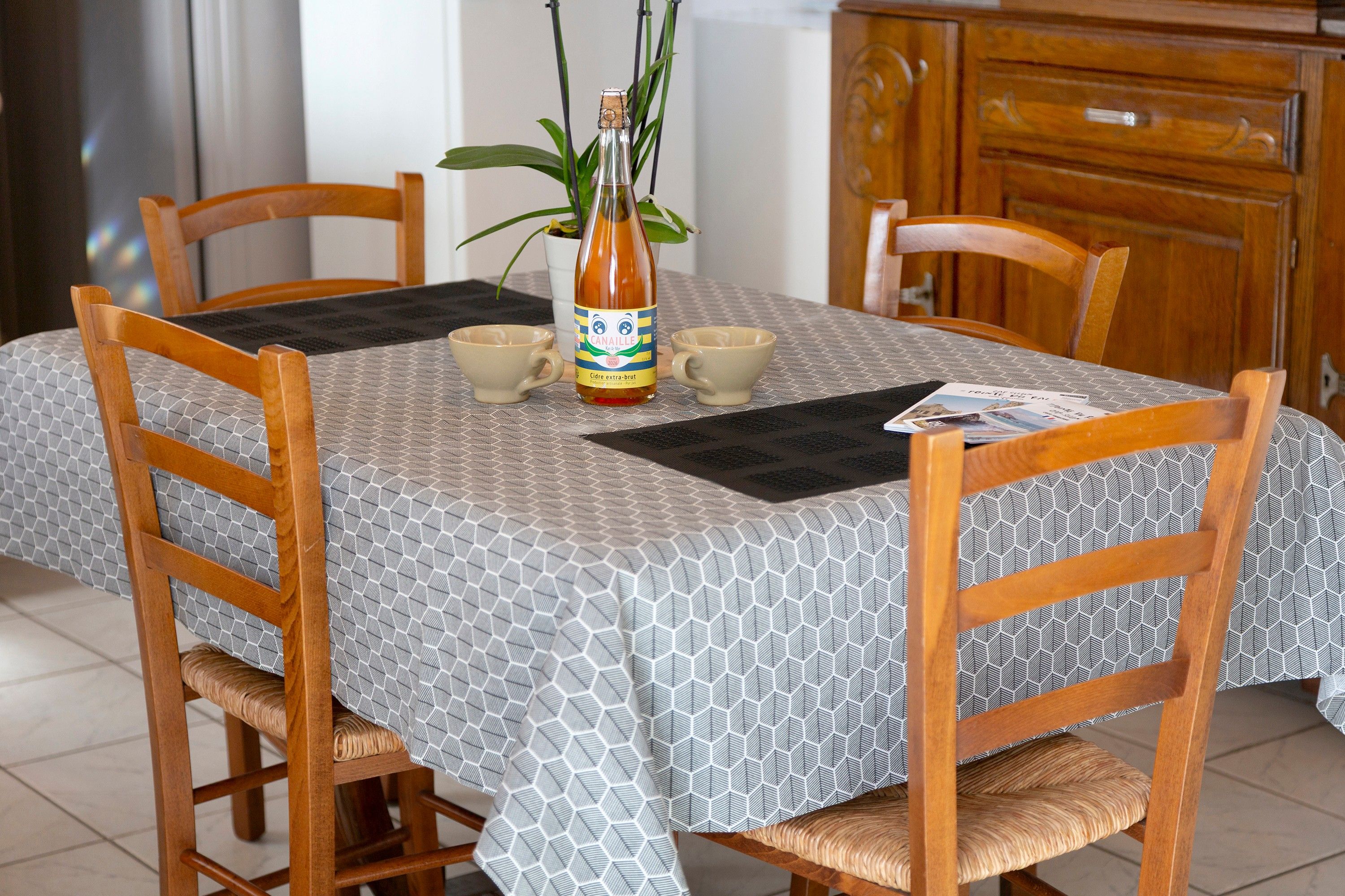 A dining table with a geometric patterned tablecloth, two wooden chairs, a bottle of Cidre extra-brut, two cups, and a plant.